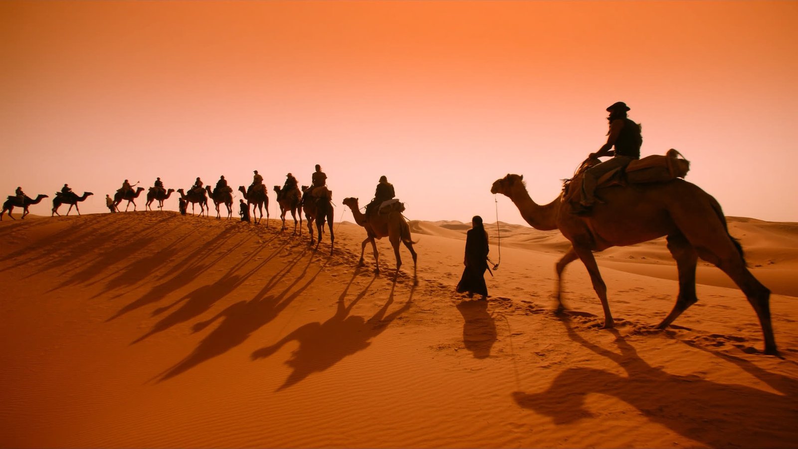 A camel caravan crossing golden Erg Chegaga dunes at sunset, riders silhouetted